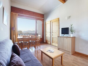 Ein Wohnzimmer mit Sofa, einem Esstisch mit Stühlen, einem Fernseher auf einem Schrank und Bergblick durch ein großes Fenster.