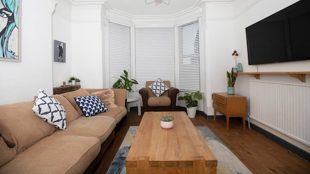 A living room with a brown sofa, a wooden coffee table, and a TV mounted on the wall. - Arthouse Lovely Spacious 5 bed house (Nottingham)