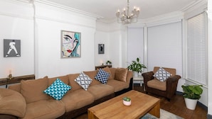 A living room with a brown sofa set, a wooden coffee table, and a chandelier. There are two potted plants and framed artwork on the walls. - Arthouse Lovely Spacious 5 bed house (Nottingham)