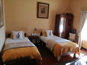 A room with two single beds, a wooden chair, a small table with a lamp, and a framed picture on the wall.