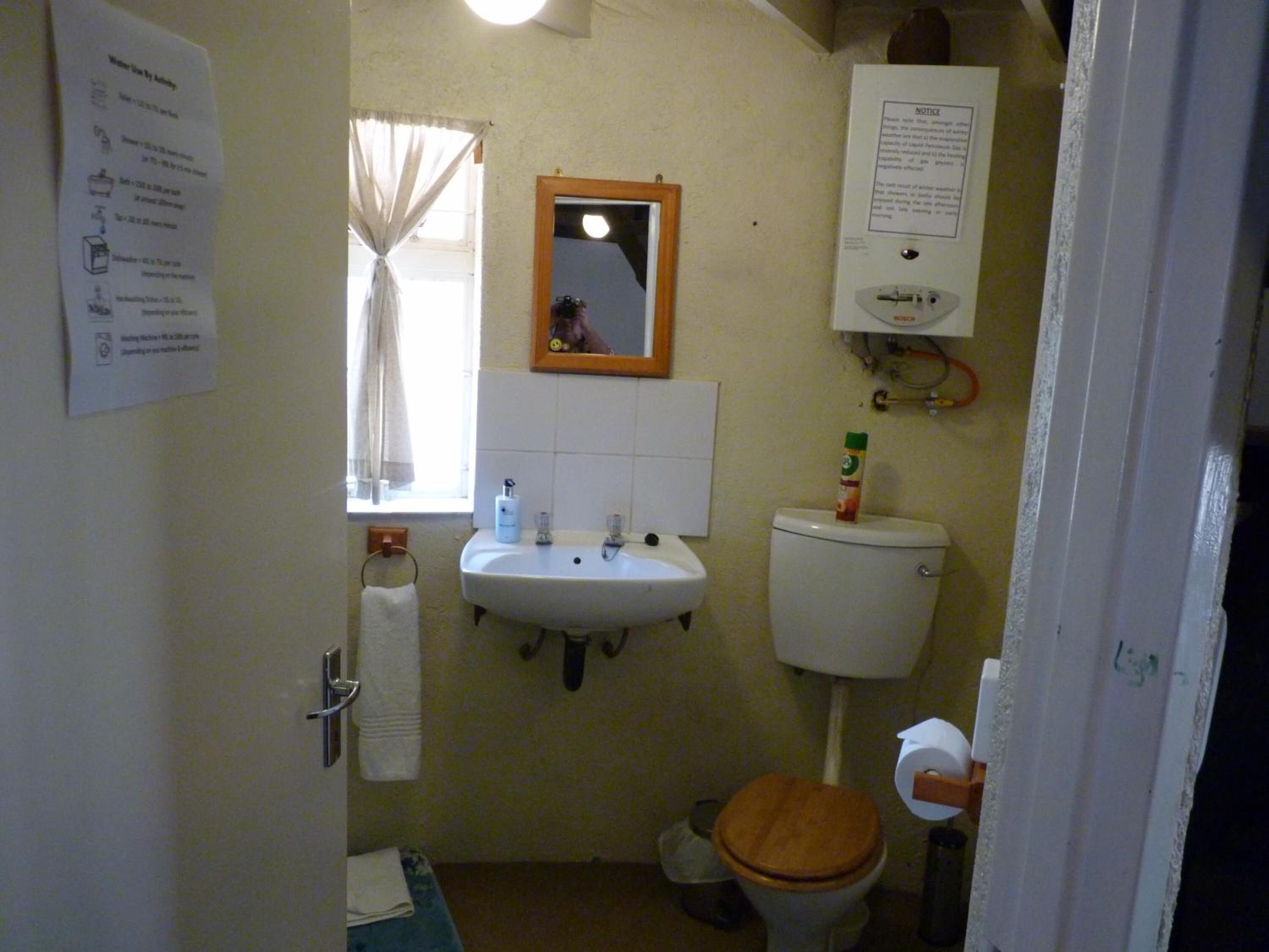 Et lille badeværelse med håndvask, toilet og spejl. Der er et vindue med gardiner, en håndklædestang og et skilt på væggen.