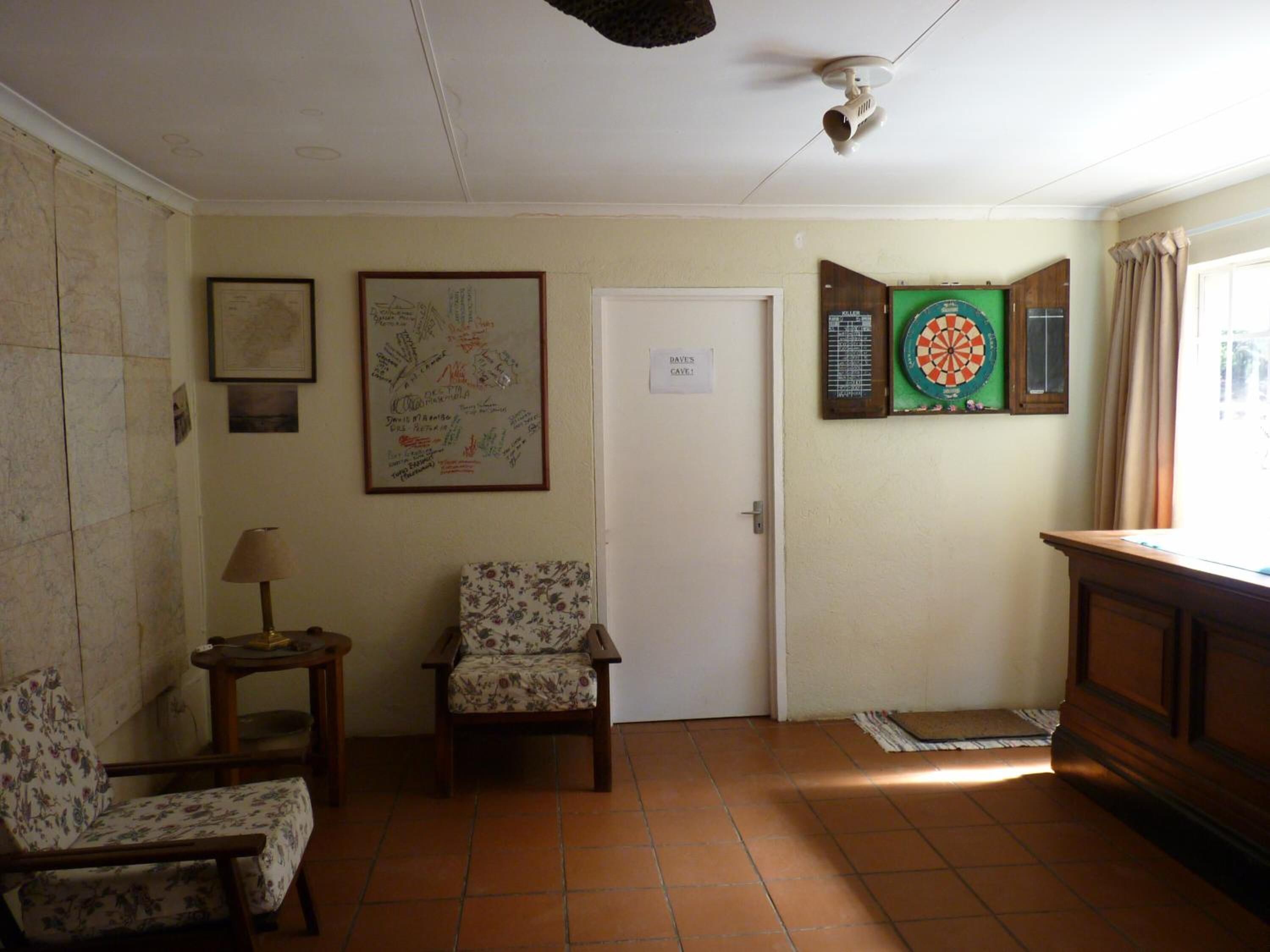 A room with a tiled floor, a door, a framed map, a dartboard, and two floral-patterned chairs.