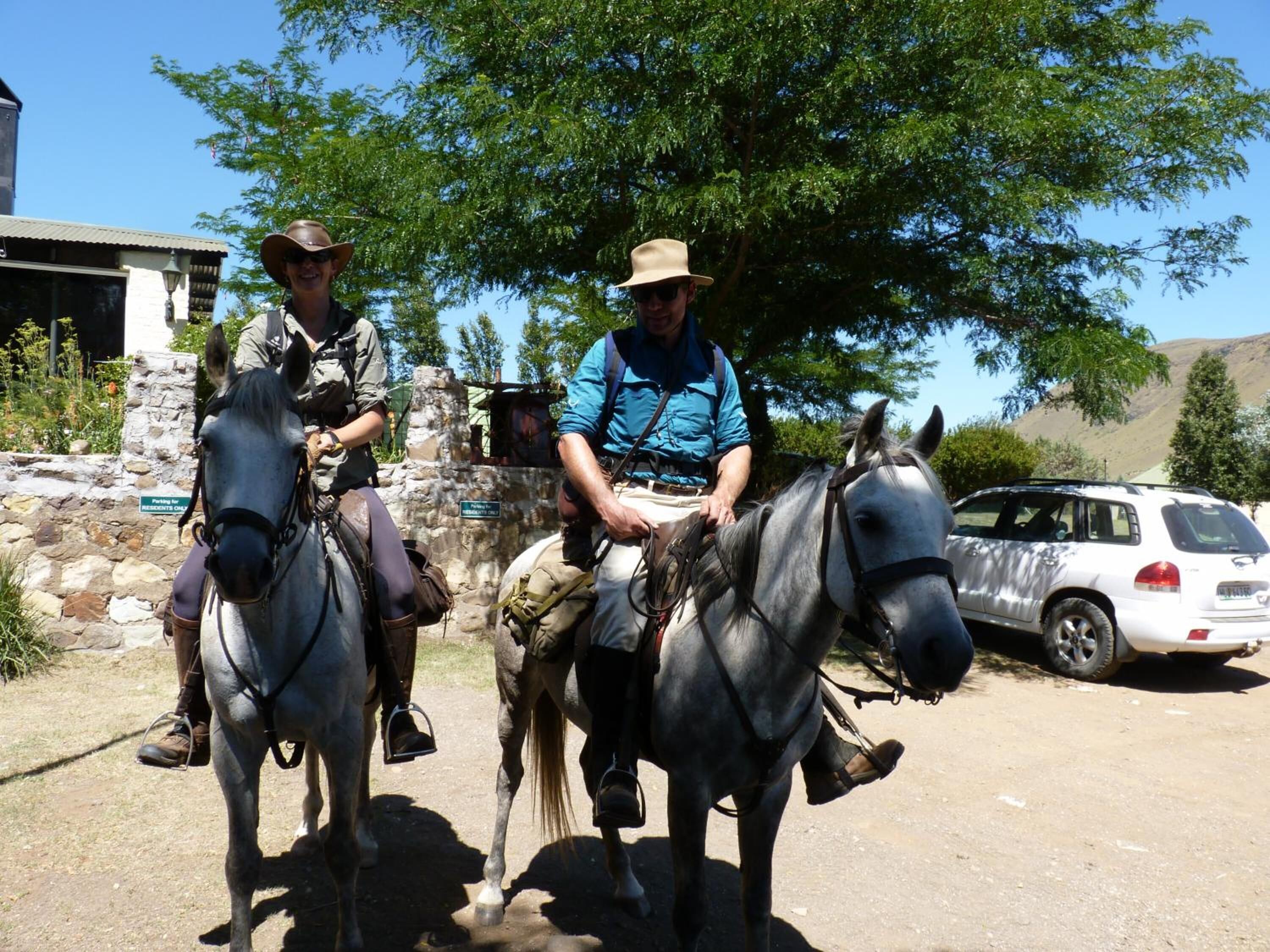 Dues persones cavalcant cavalls per un camí de terra, amb una paret de pedra i un cotxe estacionat al fons.