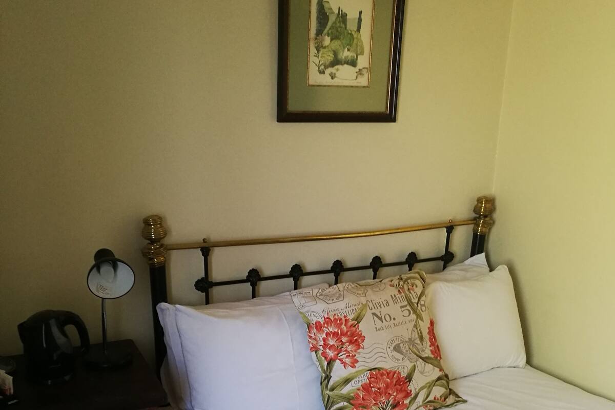 Cama bem arrumada com lençóis brancos e um cobertor vermelho, mesa de cabeceira com abajur e um quadro na parede.