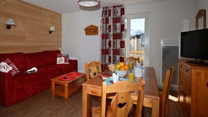 Een gezellige woonkamer met een rode bank, een houten eettafel en stoelen. Een televisie is aan de muur bevestigd, en er is een raam met uitzicht op gebouwen.