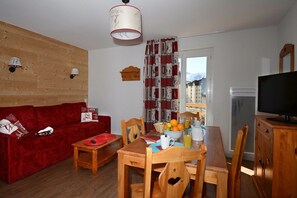 A cozy living room with a red sofa, wooden dining table, and chairs. A television is mounted on the wall, and there is a window with a view of buildings. - Apartment in France with Flexible Options (Les Deux Alpes)