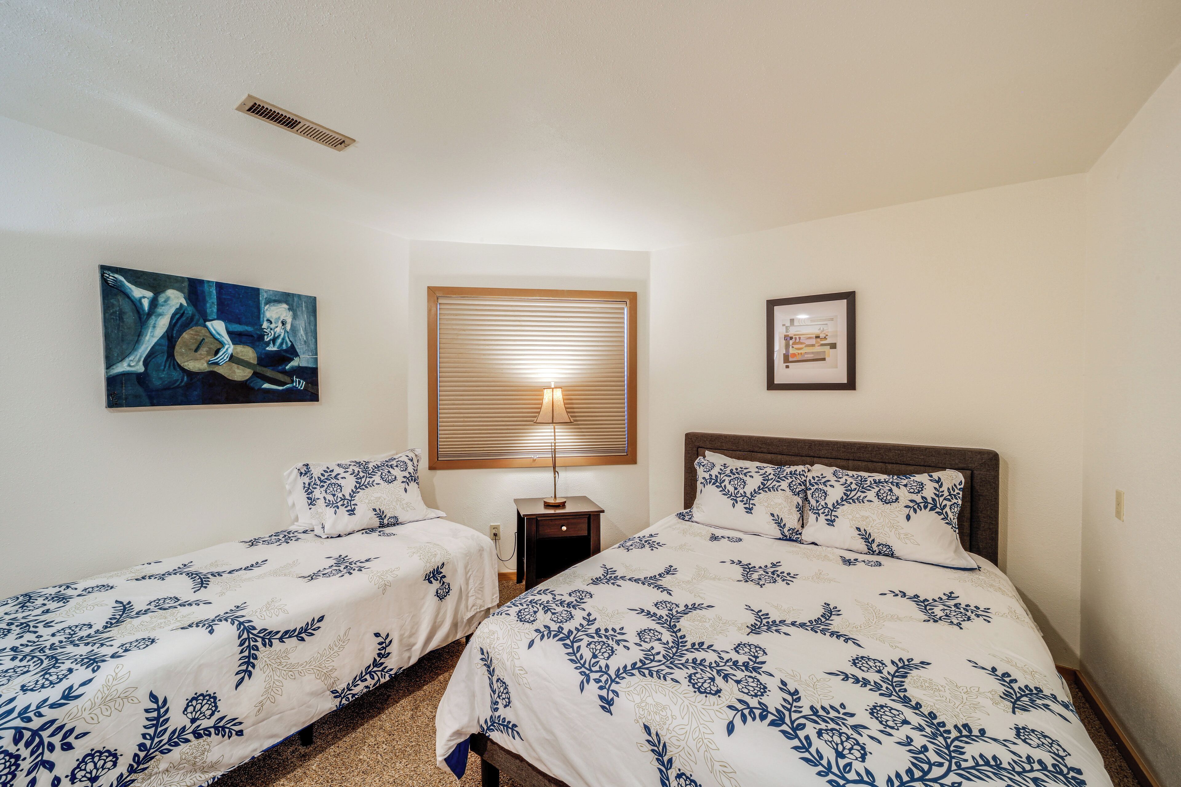 A bedroom with two beds, a window with blinds, a lamp, and a painting on the wall.