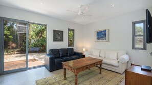 A living room with a sofa, a coffee table, and a TV. There is a sliding glass door leading to a patio with a bench. - Moffat Retreat Stylish 3BR Beach Home (MOFFAT BEACH)