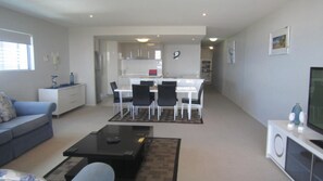 A modern living room with a sofa, coffee table, and TV stand. There is a dining area with chairs and a table. The kitchen is visible in the background with cabinets and appliances. - Outlook U33 Panoramic Ocean Views & Balcony (Kings Beach)
