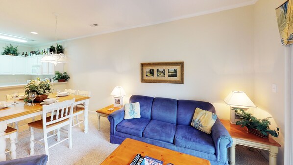 A living room with a blue sofa, a wooden coffee table, and a dining area with a white table and chairs. - Golf Villa in Little River (K102) (Little River)
