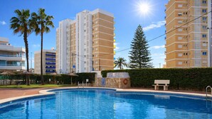 A swimming pool with a clear blue water, surrounded by tall buildings, palm trees, and a sunny sky. - Anacasa Zurich Les Marines AP3216 (Denia)