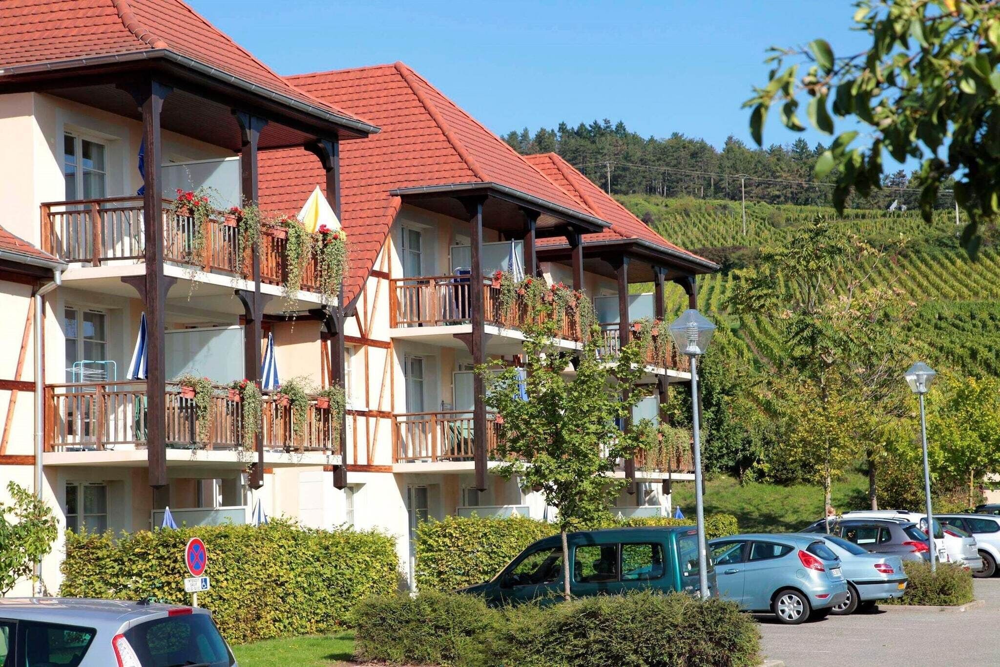 Un hotel de varios pisos con balcones, un estacionamiento y un paisaje montañoso al fondo.