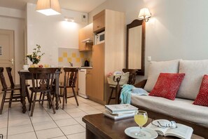 A living room with a sofa, a coffee table with books and a glass of wine, a dining area with chairs and a table, and a kitchen area with a microwave.