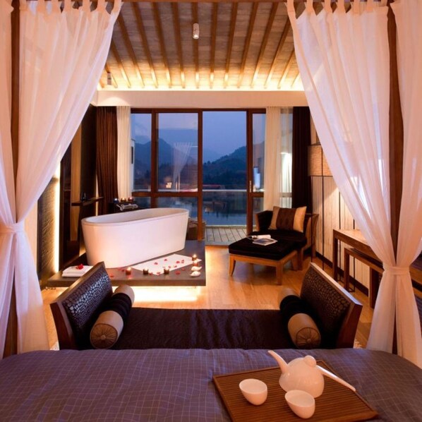 A bedroom with a large bed, a sitting area with a table and chairs, a bathtub, and a view of mountains through large windows.