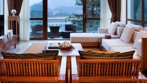 Un salon moderne doté d’une grande fenêtre donnant sur les montagnes, d’un canapé, de deux fauteuils en bois et d’une petite table sur laquelle est posé un vase de fleurs.