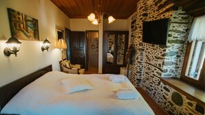 A bedroom with a large bed, stone wall, wooden ceiling, and a TV mounted on the wall. - Eudora - Mansion (Zagora-Mouresi)