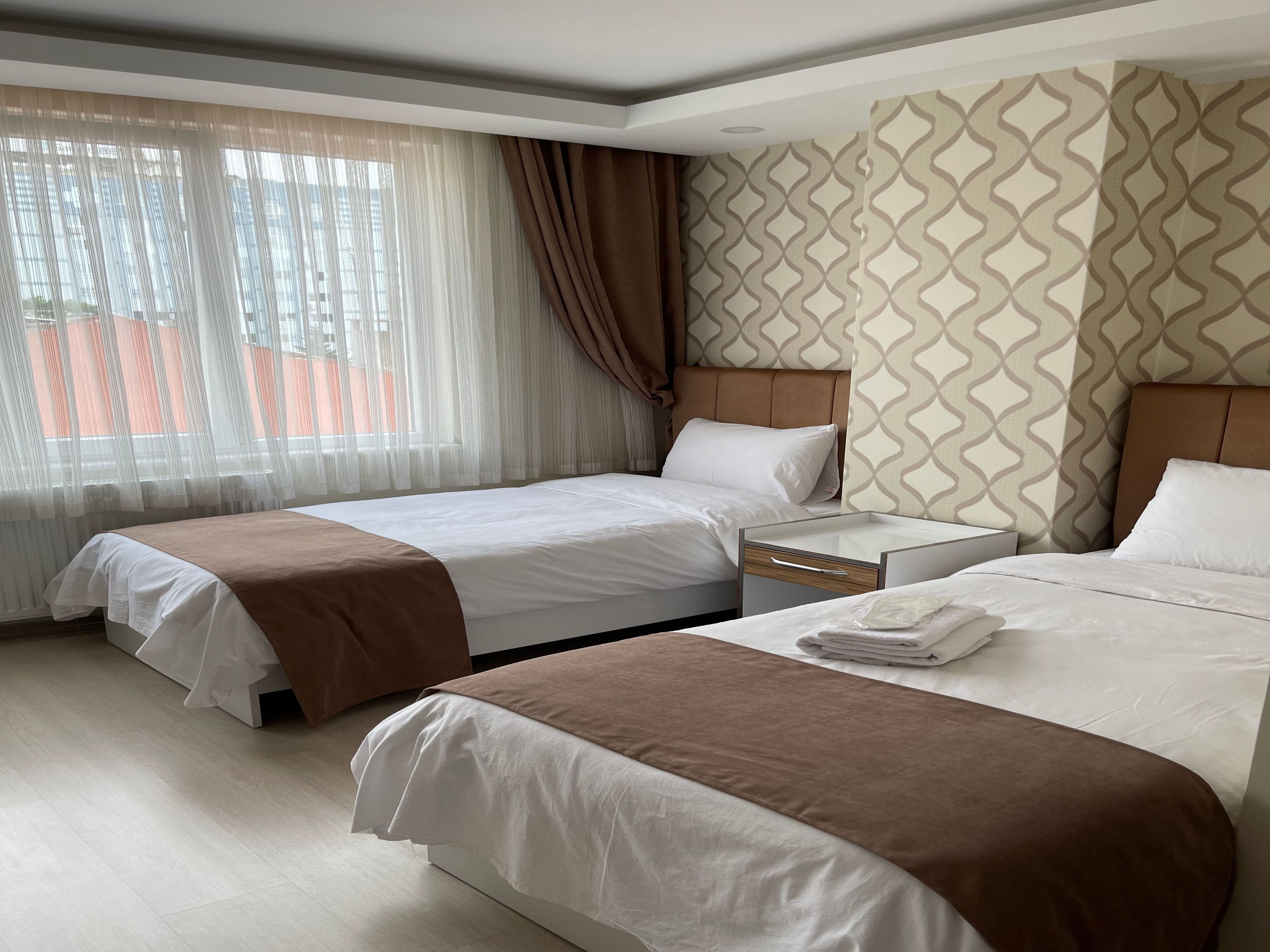 Ein Hotelzimmer mit zwei Betten, einem Nachttisch, einem Fenster mit durchscheinenden Vorhängen und einer Wand mit Muster.