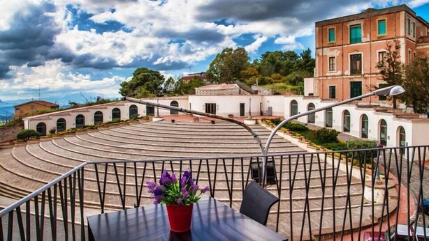 A terrace with a curved seating area, a potted plant, and a view of a historic building with arched windows. - B&B La Casa dei Miraglia San Cataldo (San Cataldo)