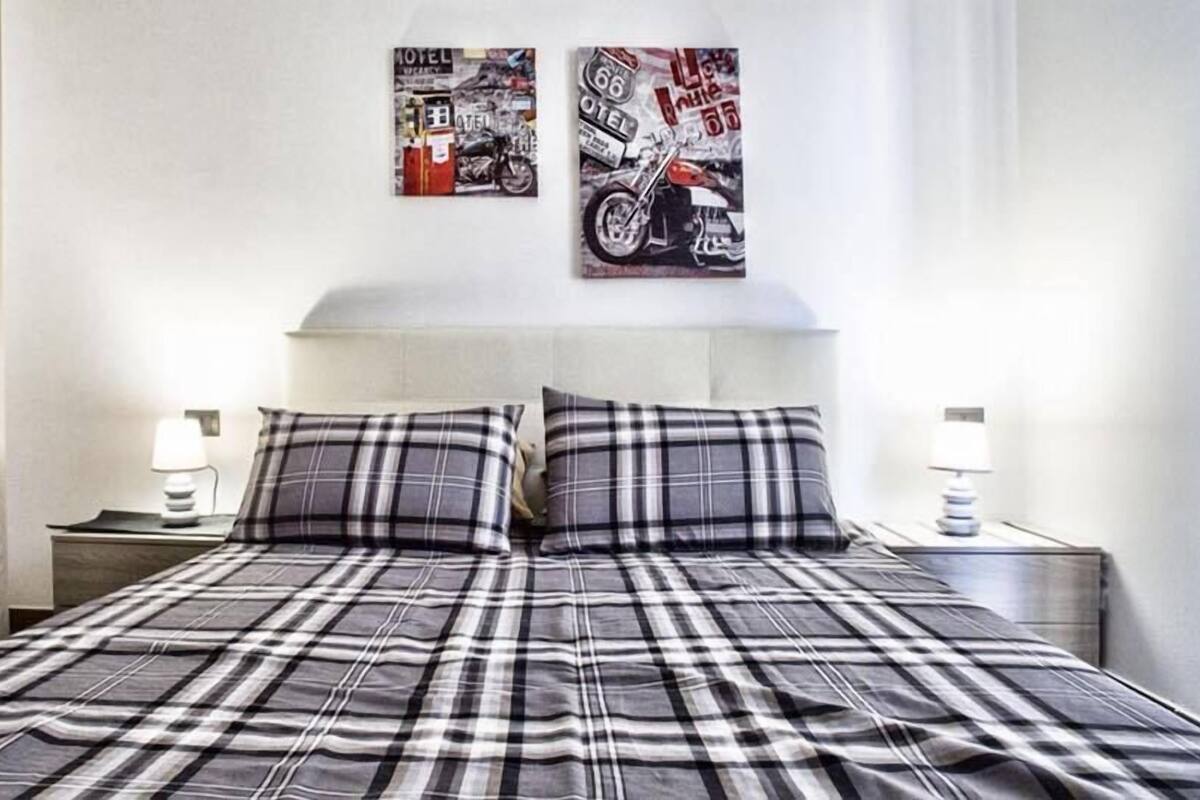 Ein ordentlich bezogenes Bett mit einer Karo-Decke, zwei Nachttischlampen und zwei Wandbildern mit Motorrädern.