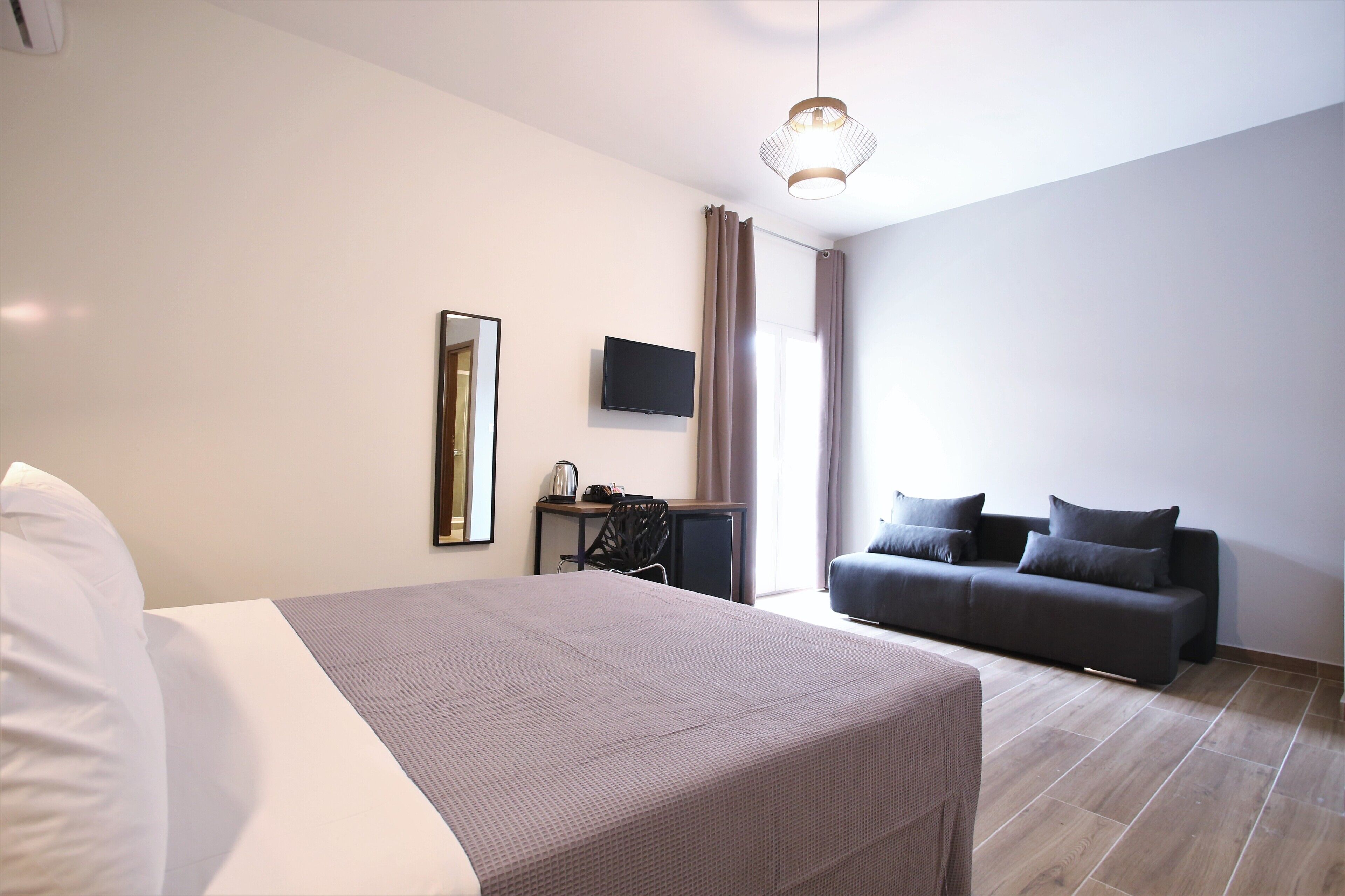 A hotel room with a bed, a desk with a chair, a TV, a sofa, and a large window with curtains.