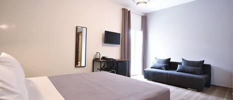 A hotel room with a bed, a desk with a chair, a TV, a sofa, and a large window with curtains.