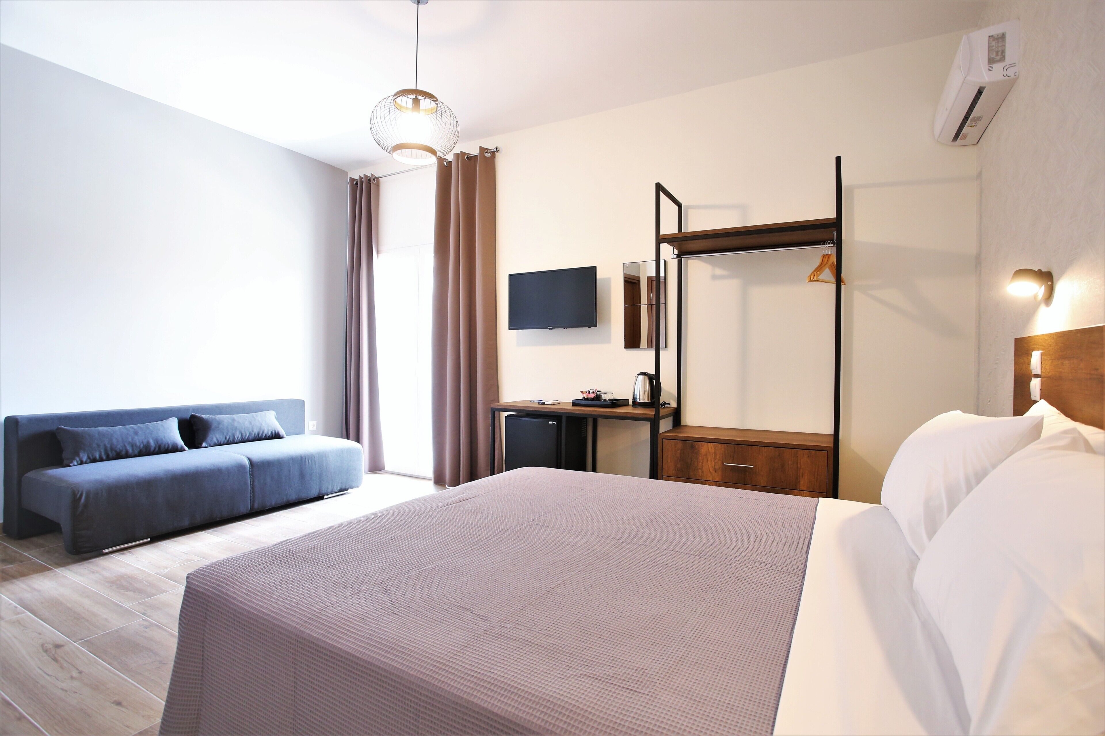 A modern hotel room with a large bed, a sofa, a desk with a flat-screen TV, and a wardrobe.
