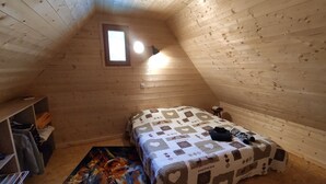 A small, sloped-roofed room with wooden walls, a single bed with a quilt, a small table, and a shelf with items.