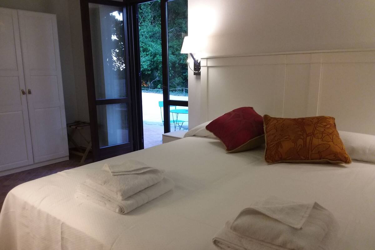 Un lit bien fait, avec deux oreillers, une tête de lit blanche et une vue sur un espace extérieur grâce à une porte vitrée.