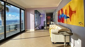 Un intérieur moderne avec une grande fenêtre donnant sur la mer, un canapé blanc et une peinture abstraite aux couleurs vives accrochée au mur.