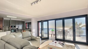 Un salon moderne avec un canapé gris, une table basse et une vue sur la mer grâce à de grandes portes-fenêtres coulissantes.