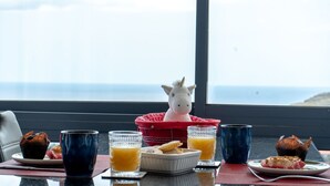 Een ontbijttafel met een eenhoornbeeldje, twee glaasjes sinaasappelsap, een bord gebakjes en een toetje, alles op een tafel bij een raam met uitzicht op de zee.
