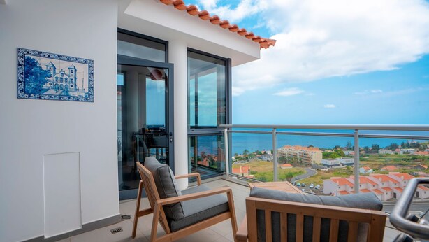 Een balkon met uitzicht op zee en gebouwen, een betegelde muur met een blauw-wit ontwerp, en een houten stoel en tafel.