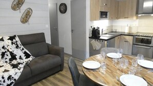 A compact living space with a sofa, a dining table set for four, a kitchenette, and a clock on the wall. - Charming 2-Bedroom Apt with Balcony, Mountain Views, WiFi, Parking & Baby Gear in St Lary Soulan (Vignec)