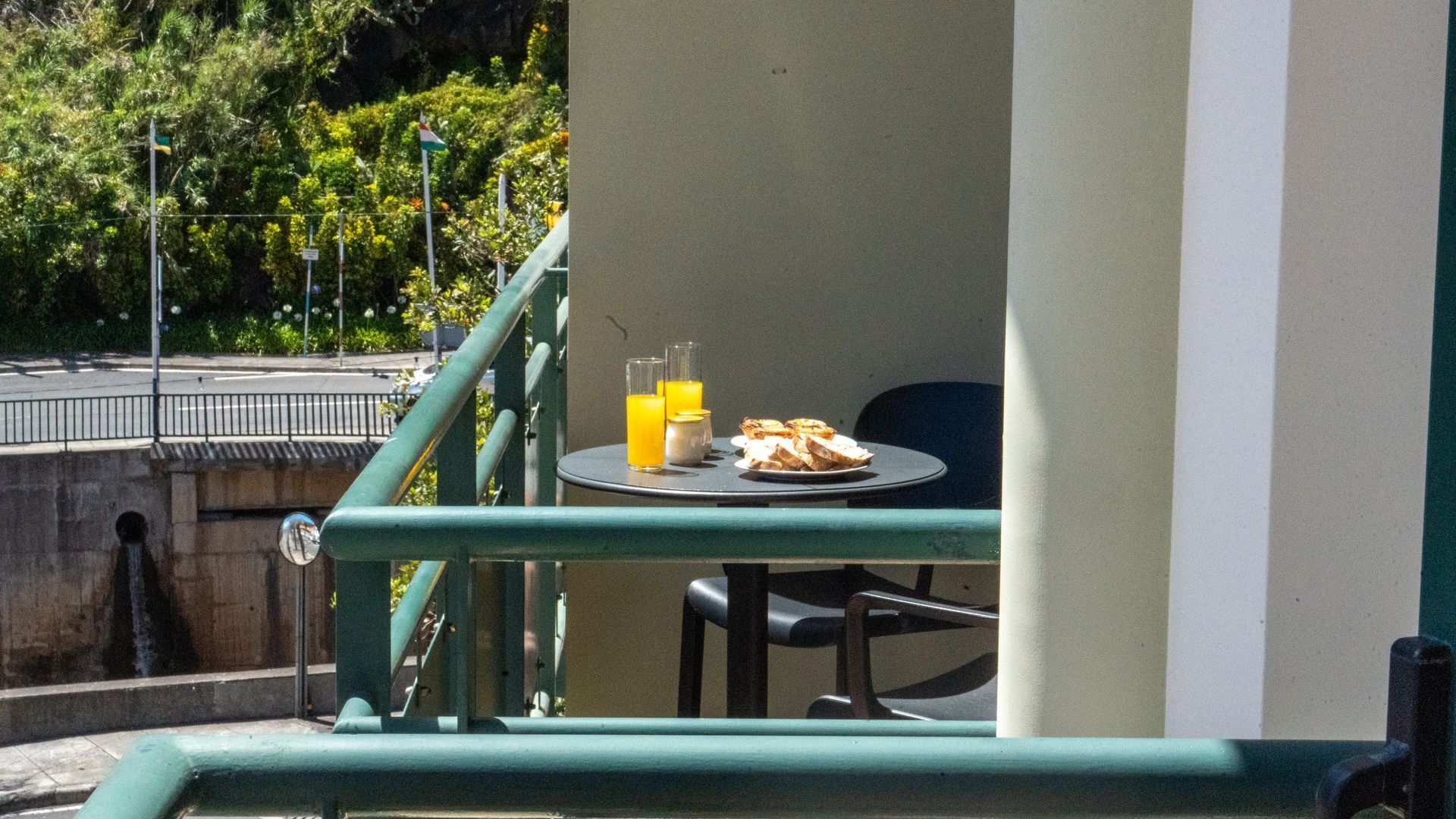 A balcony with a table set for breakfast, including toast and orange juice, overlooking a street with greenery and a fence.