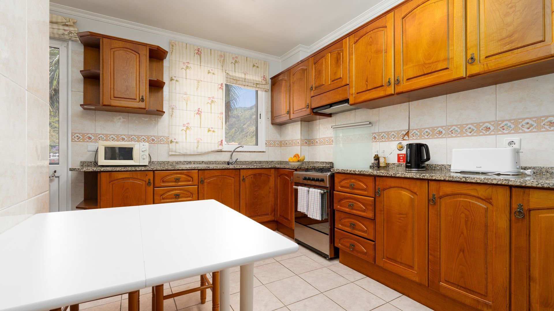 A kitchen with wooden cabinets, a white countertop, a stove, a microwave, and a window with a curtain.