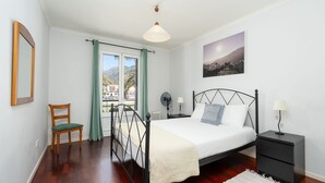 Une chambre à coucher comprenant un lit, une chaise, une table de chevet, une lampe, un tableau au mur et une fenêtre avec des rideaux.