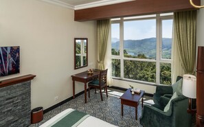 A hotel room with a bed, a desk, a chair, a sofa, and a large window offering a scenic view. - Fort Munnar (Devikolam)