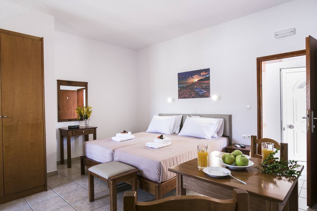 A hotel room with two beds, a wooden wardrobe, a bedside table with a vase of flowers, a painting on the wall, and a breakfast table with apples and juice.