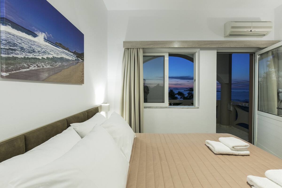 A hotel room with a bed, a window with a view of the beach and sunset, and a balcony with a glass door.