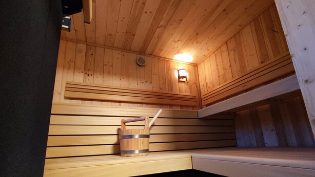 Eine Sauna mit Holzwänden und einer Sitzbank. Ein Eimer mit Tragegriff und eine Wandleuchte sind an der Wand angebracht.