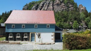 Ein traditionelles blaues Haus mit rotem Dach, gelegen in ländlicher Umgebung mit einem felsigen Berg im Hintergrund.
