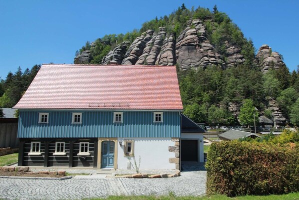 A traditional blue house with a red roof, situated in a rural area with a rocky mountain in the background.