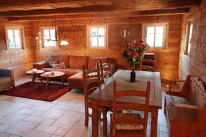 A cozy log cabin interior with a wooden dining table, chairs, a sofa, and a red rug.