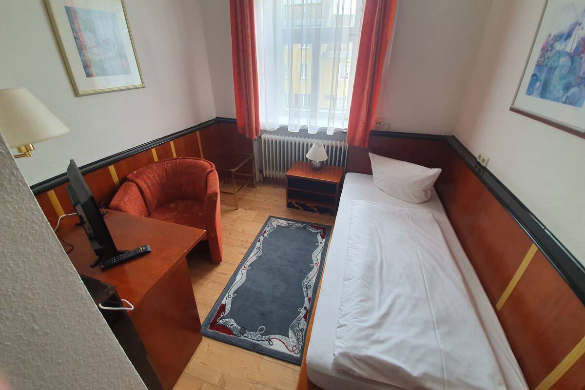 A hotel room with a bed, a desk, a chair, a radiator, and a window with curtains.