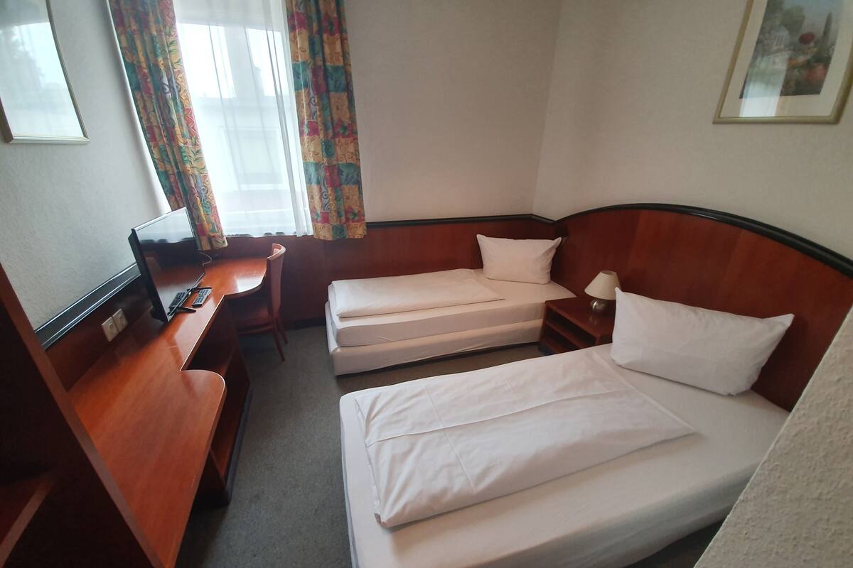 A hotel room with a wooden desk, a television, two single beds, and a small bedside table with a lamp.
