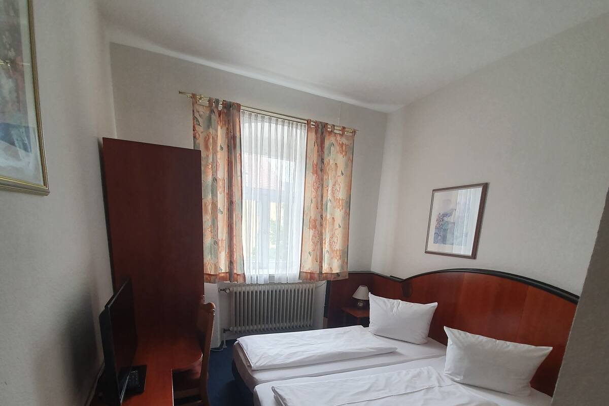 A hotel room with two beds, a desk, a chair, a TV, a radiator, and a window with curtains.