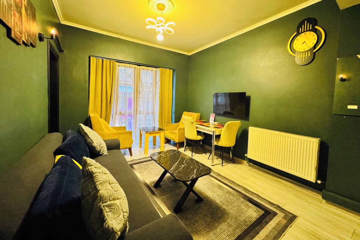 A living room with a sofa, chairs, a coffee table, a TV, a clock, and a window with curtains.
