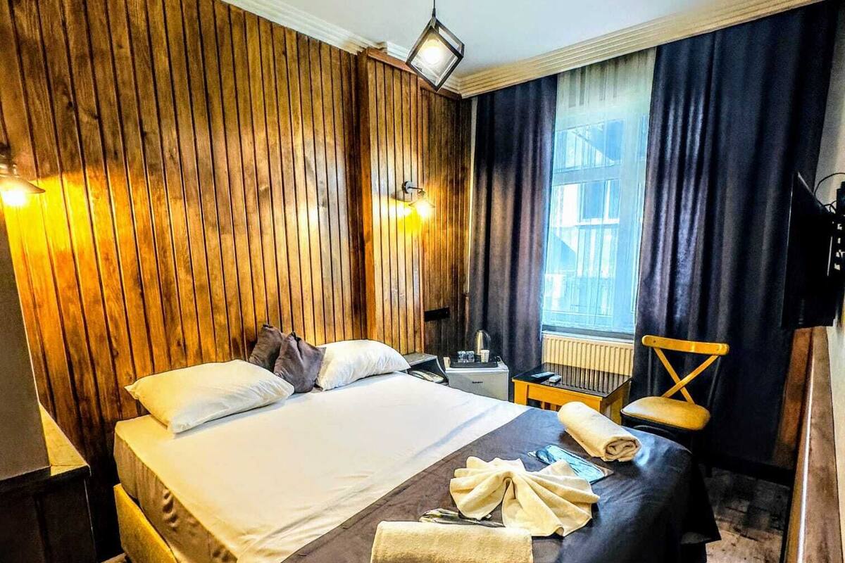 A hotel room with a wooden panel wall, a bed with white linens, a desk with a lamp, and a window with curtains.