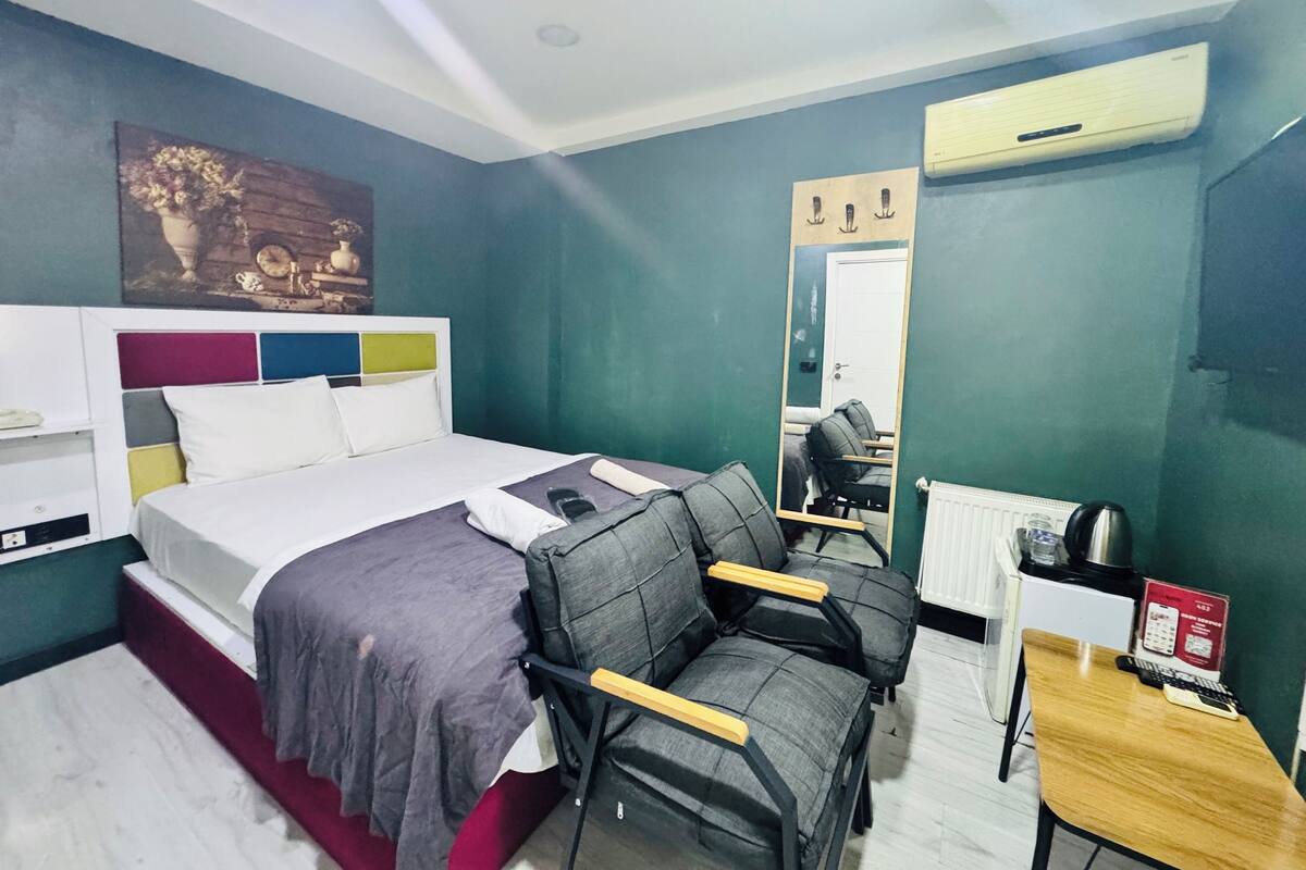A hotel room with a bed, two chairs, a desk, a mirror, and an air conditioning unit.