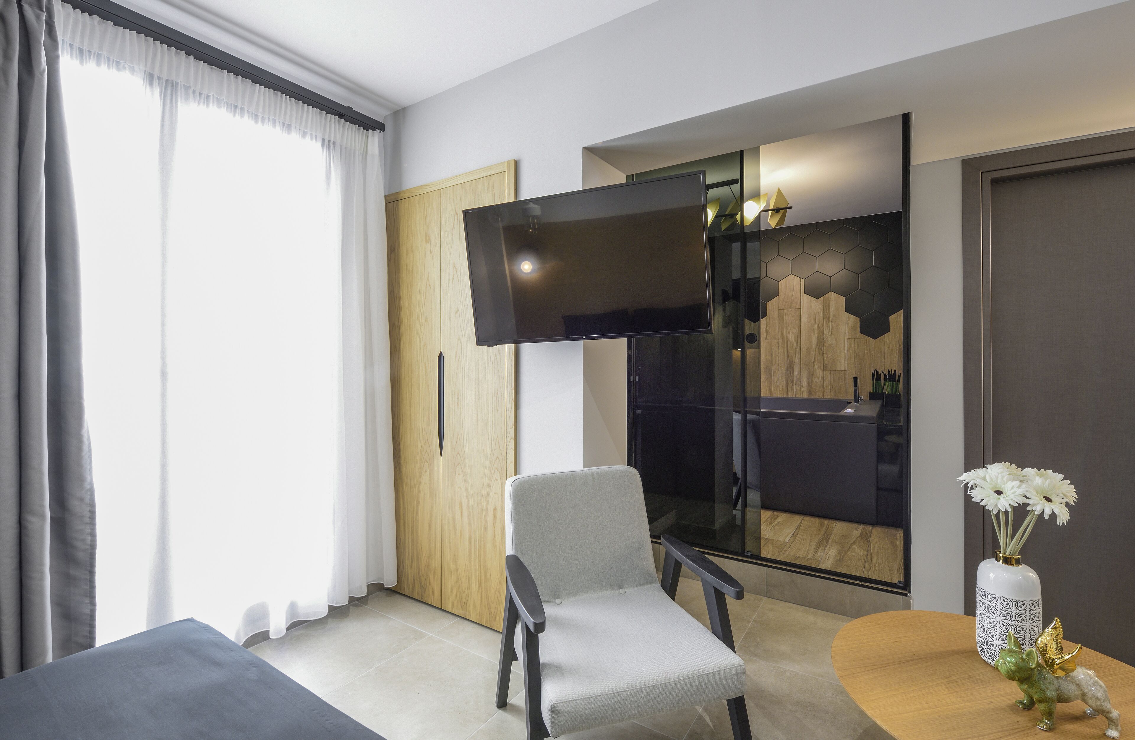 Una camera d'albergo moderna con una grande televisione a schermo piatto, una poltrona grigia, una porta in legno e un vaso con fiori su un tavolo rotondo.